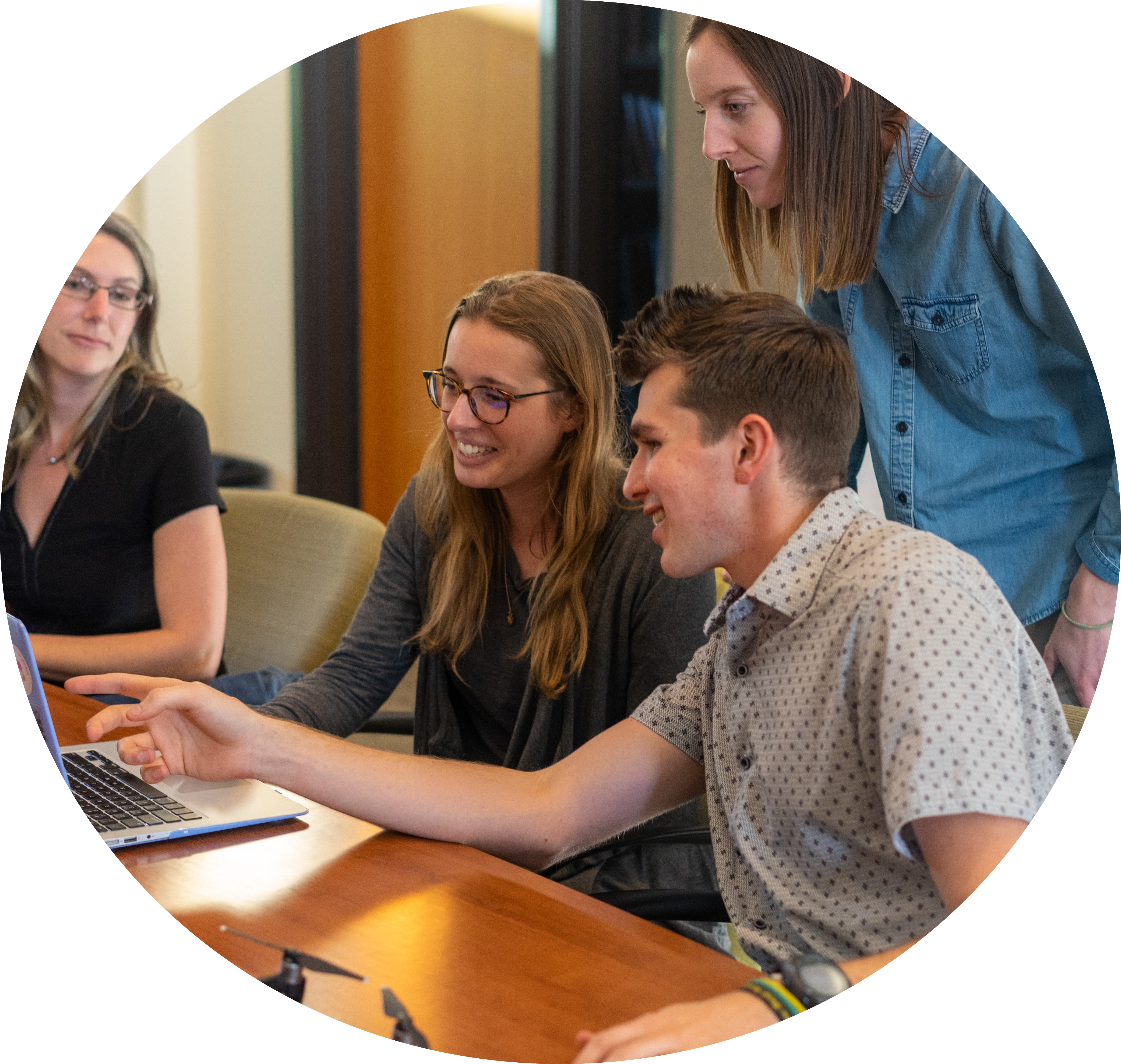 Multiple smiling people at a desk are crowded around a laptop