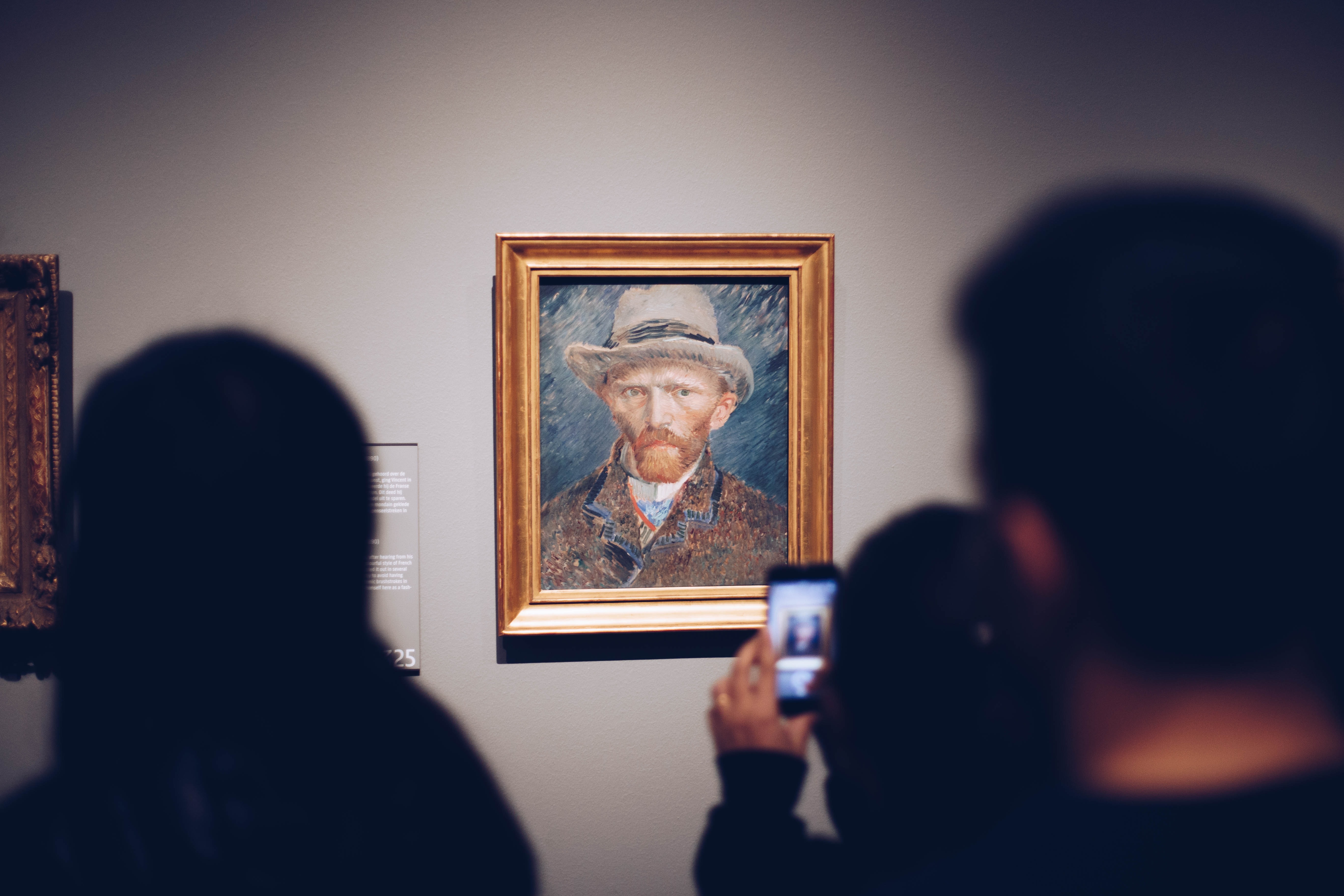 Van Gogh's 'Self Portrait with a Grey Felt Hat' is seen on the gray wall of a museum in a gold frame. In it, the artist is pictured from the shoulders-up, looking slightly to the left with a stoic gaze, while wearing a brown coat. Some blurry museum visitors are in the foreground, with one taking a picture of the artwork on her cell phone.